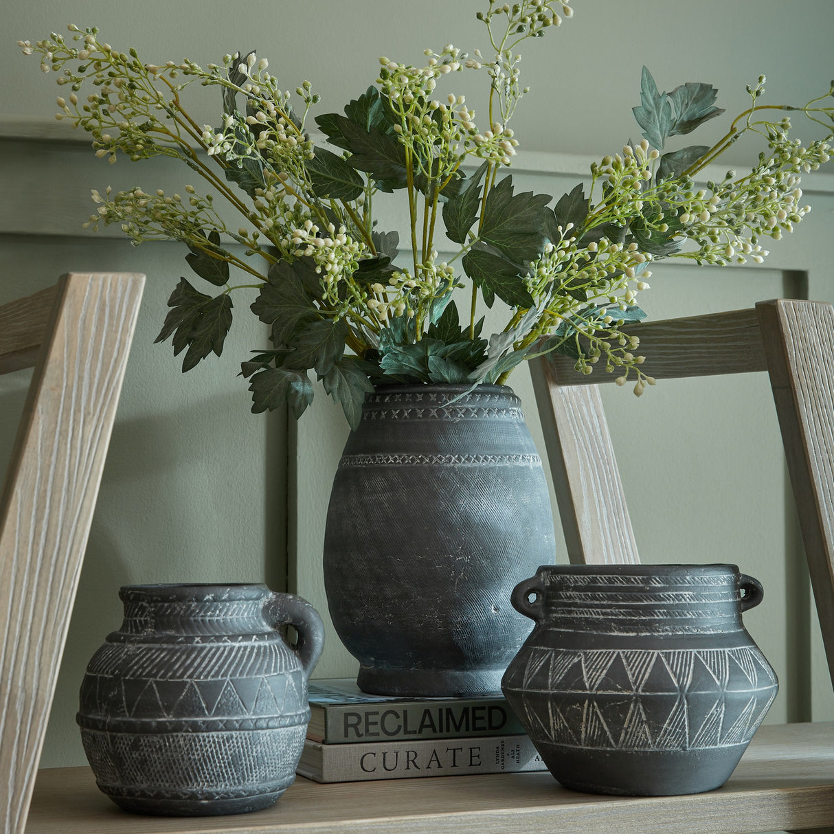 Hill Interiors Milk Thistle Bud Spray displayed with foliage in a dark patterned ceramic vase, flanked by two smaller tribal-style pots on styling books.
