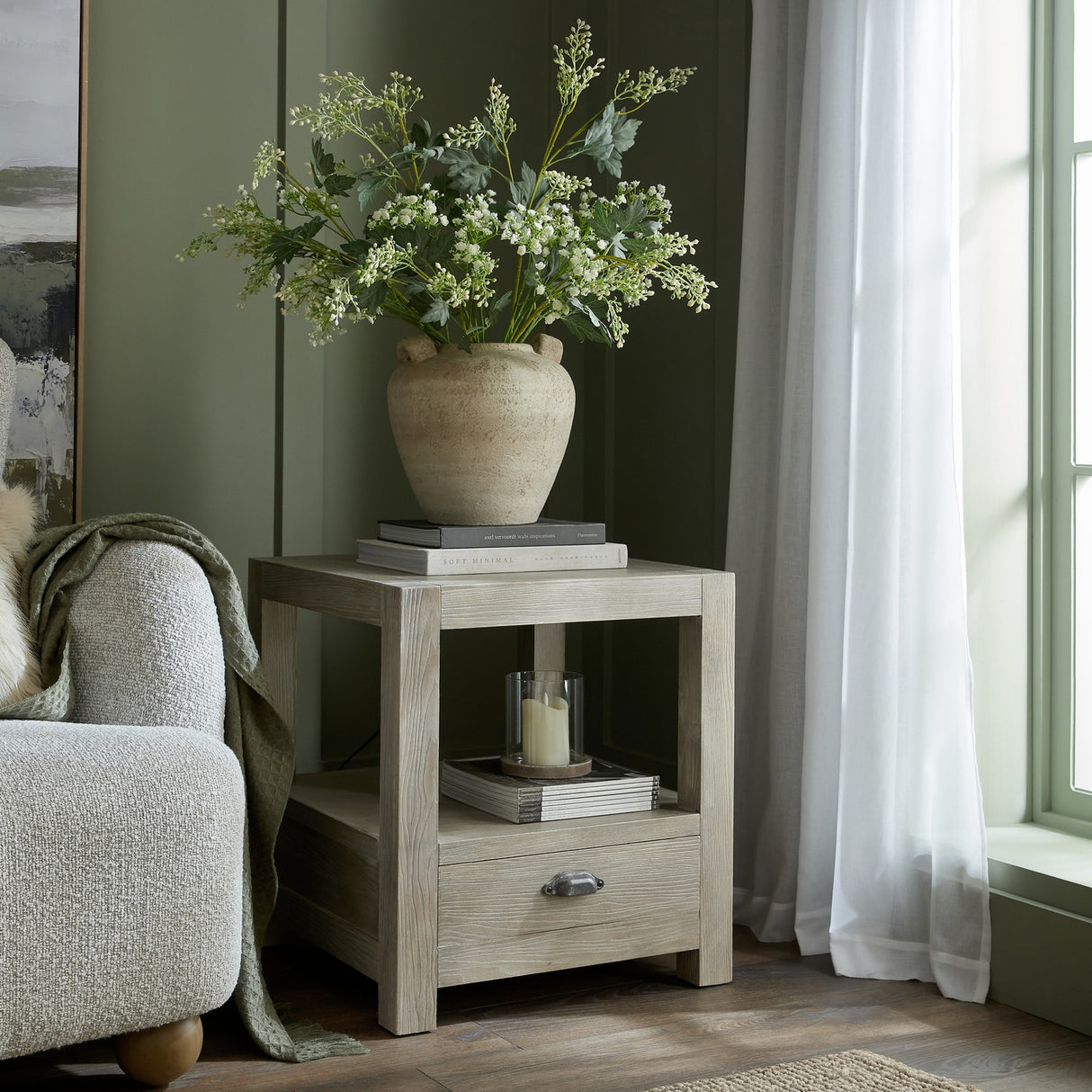 Hill Interiors Milk Thistle Bud Spray arranged in a textured stone vase atop books on a washed wood side table in a sage green living room.