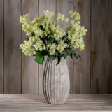 Hill Interiors Green French Meadow Spray with pale green blooms and leafy foliage arranged in a ribbed stone vase against a wooden plank backdrop.