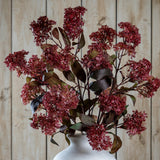 Hill Interiors Purple French Meadow Spray with clustered deep red-purple blooms and green-brown foliage arranged in a white ceramic vase against wood panelling.