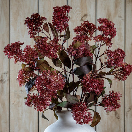 Hill Interiors Purple French Meadow Spray with clustered deep red-purple blooms and green-brown foliage arranged in a white ceramic vase against wood panelling.