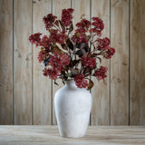 Hill Interiors Purple French Meadow Spray with clusters of deep pink-purple blooms and dark foliage arranged in a stone ceramic vase against wood panelling.