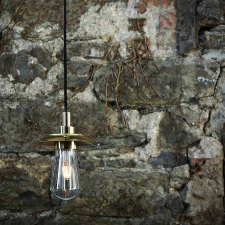 Ren Glass Disc Pendant Light IP65 in polished brass with a flat disc hood over a teardrop glass shade and visible filament bulb, shown against a rustic stone wall.
