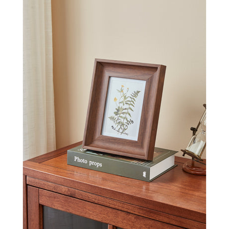 Renabie Photo Frame 21 x 26cm in a wood-effect finish with wide bevelled moulding, displaying a botanical print, standing on a book atop a wooden sideboard.
