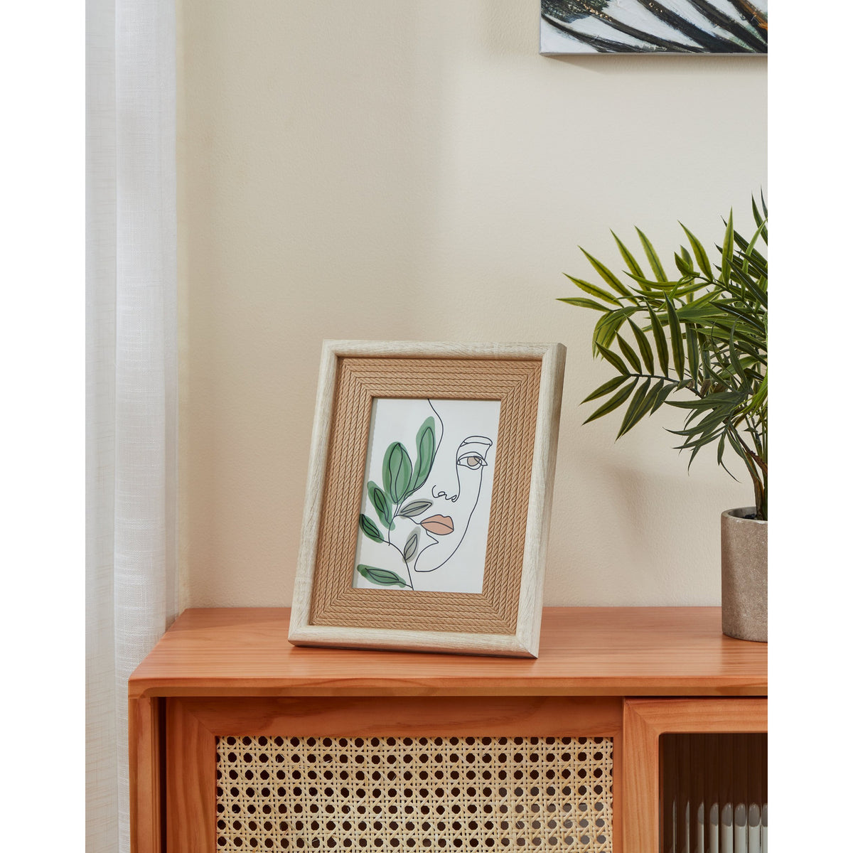 Renabie Photo Frame 22 x 27cm in wood-effect finish with rope-textured inner border, displayed on a wooden sideboard beside a potted plant, holding a line-art face and leaf print.