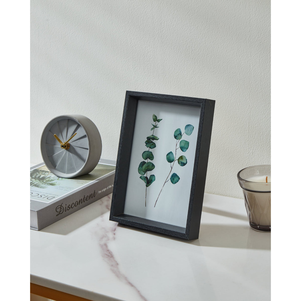 Renabie Photo Frame in black, deep box profile with glass front displaying pressed eucalyptus leaves, shown on a marble shelf beside a clock, book, and candle.