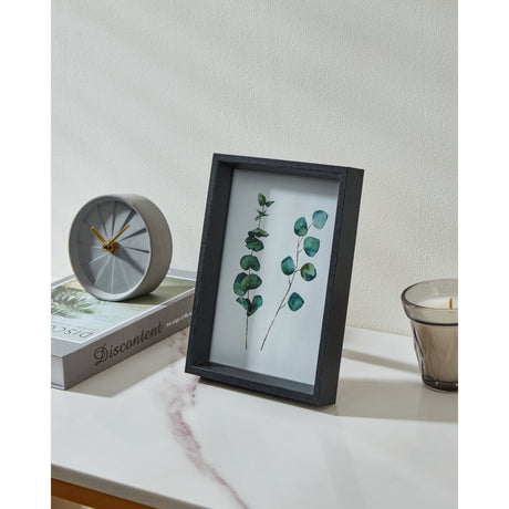 Renabie Photo Frame in black, deep box profile with glass front displaying pressed eucalyptus leaves, shown on a marble shelf beside a clock, book, and candle.