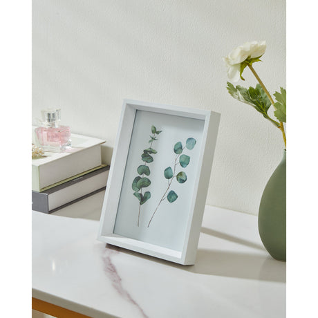 Renabie Photo Frame in white, a box-style MDF frame with glass front displaying a botanical print, shown on a marble surface beside a vase and perfume bottles.