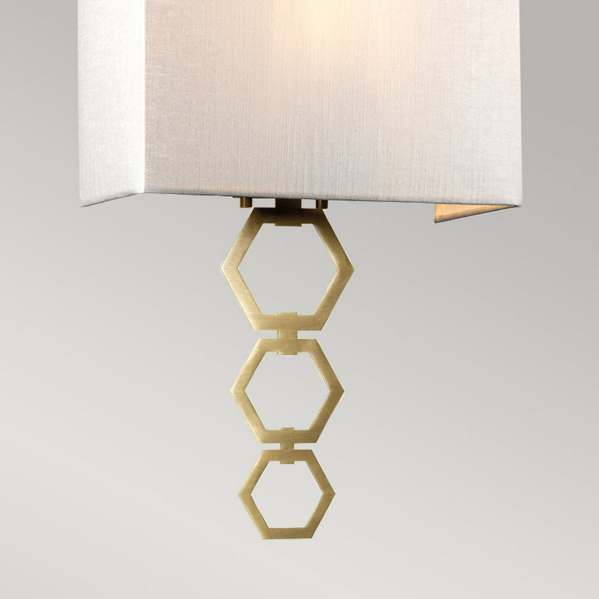 The Ria Medium 1 Light Wall Light in aged brass features an ivory faux silk shade that is suspended from the ceiling. Beneath the shade, three gold hexagonal rings are vertically connected as a decorative element, while an octagonal finial enhances its elegance against a plain, light gray background.