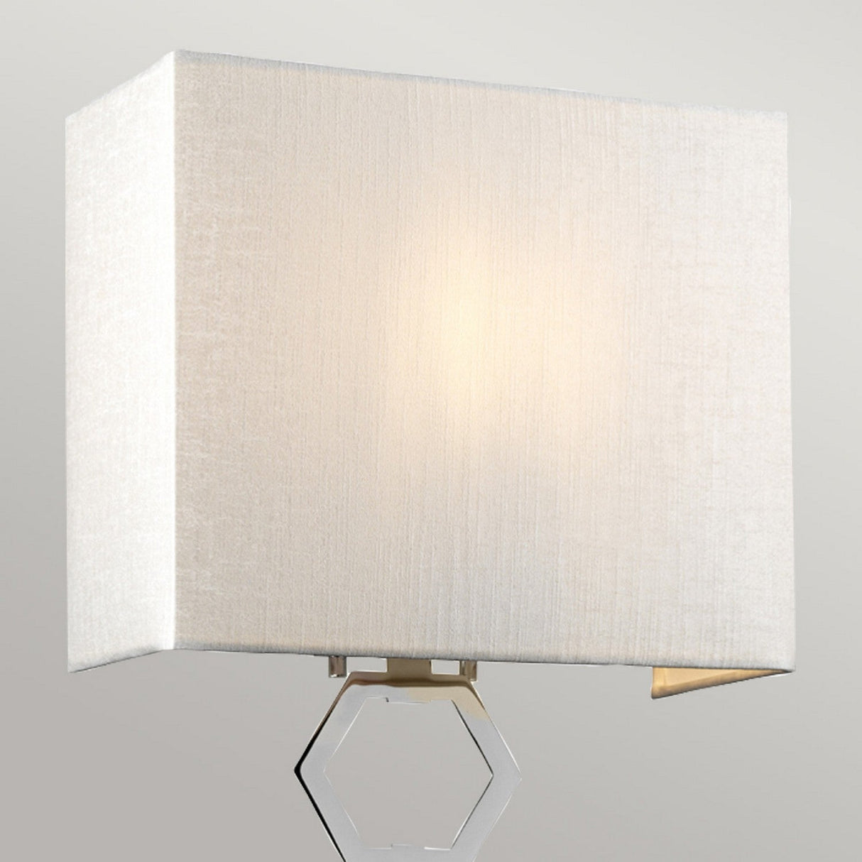 The Ria Medium 1 Light Wall Light in polished chrome showcases a beige, cube-shaped faux silk shade that casts a warm, soft glow. It is accented with a hexagonal metal detail below the shade, set against a plain light gray background.