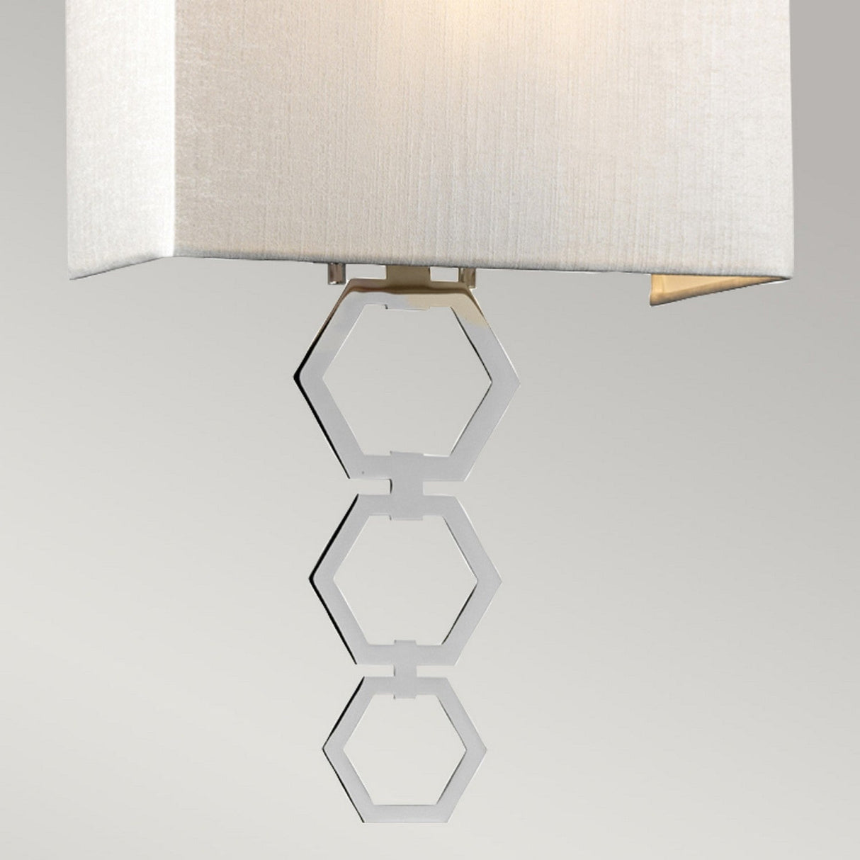 The Ria Medium 1 Light Wall Light showcases a contemporary design with a beige faux silk shade and ornamental metal hexagons suspended beneath it, all highlighted by polished chrome accents, against a simple light gray backdrop.