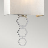 The Ria Medium 1 Light Wall Light showcases a contemporary design with a beige faux silk shade and ornamental metal hexagons suspended beneath it, all highlighted by polished chrome accents, against a simple light gray backdrop.