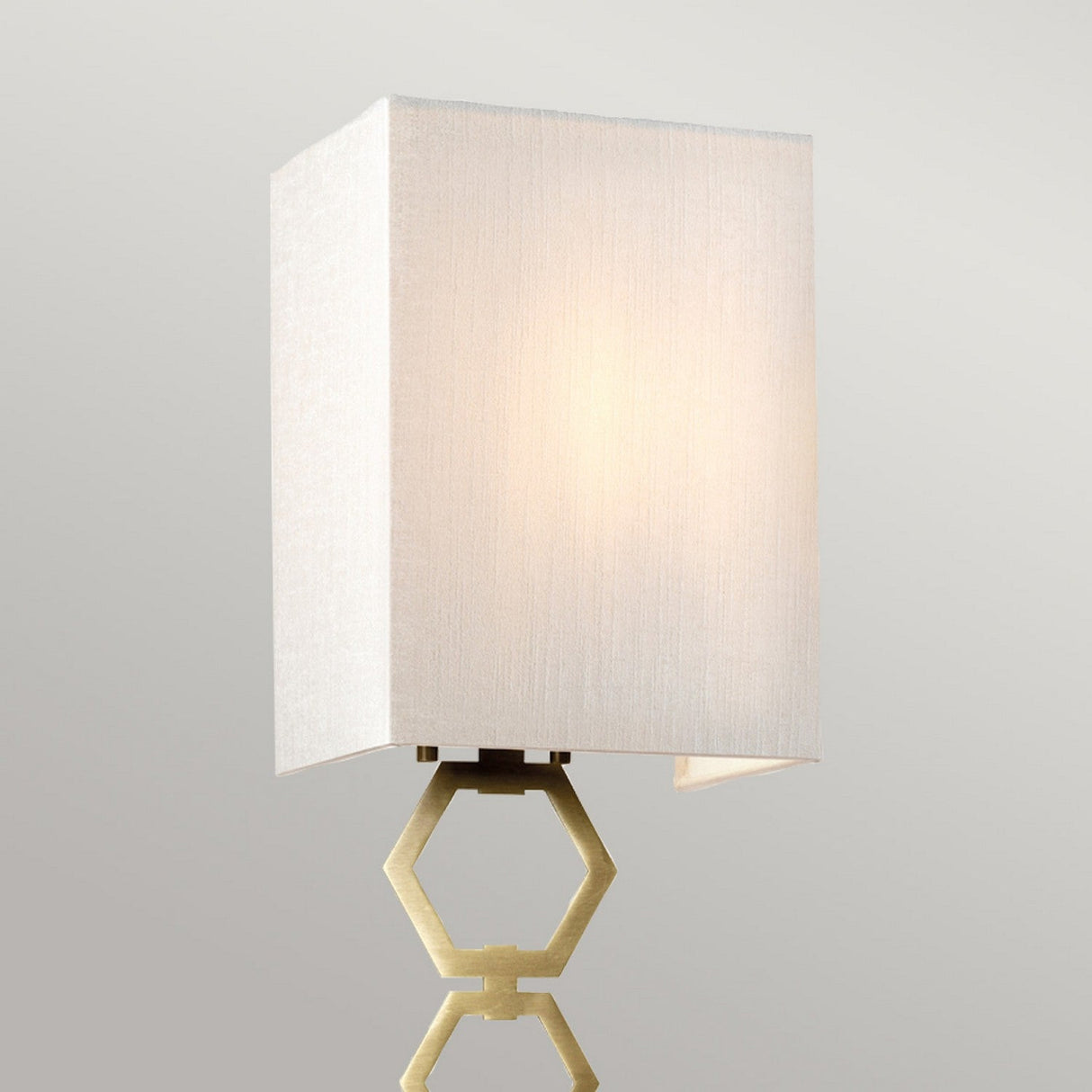 Introducing the Ria Small 1 Light Wall Light in aged brass, featuring a geometric hexagonal-shaped metallic design. It comes with an ivory faux silk shade that emits a soft glow, creating a warm and elegant ambiance.