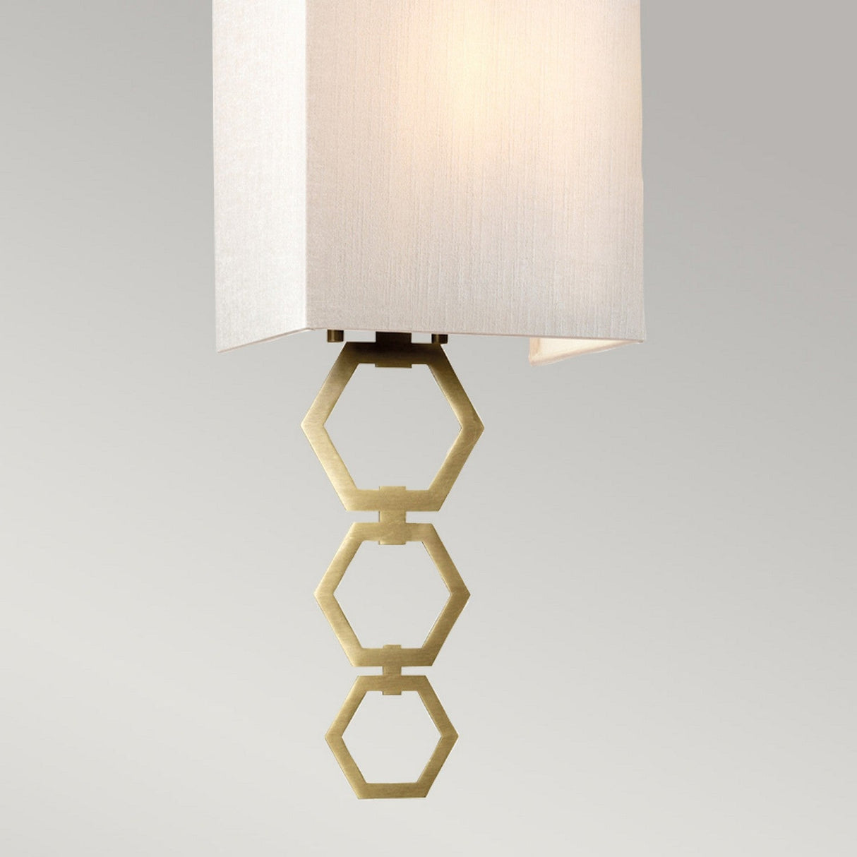 Suspended from the ceiling, the Ria Small 1 Light Wall Light - Aged Brass features a sophisticated ivory faux silk shade. Beneath this shade, an eye-catching design of three linked hexagonal rings in aged brass forms a stylish accent against a neutral grey backdrop.