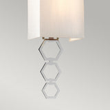 The Ria Small 1 Light Wall Light in polished chrome showcases a white rectangular shade complemented by three metallic hexagonal rings, hanging vertically to form a chic geometric accent against a neutral background.