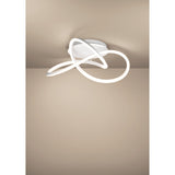Riaza LED ceiling light in white, featuring a looped ribbon-style fixture crossing over itself beneath a round ceiling canopy, mounted close to a neutral-toned ceiling.
