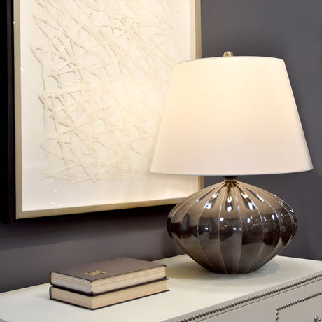 A contemporary brown and black ceramic table lamp, known as the Rib Pumpkin 1 Light Table Lamp, with a ribbed design and a faux silk shade, is placed on a light-colored dresser. Two closed books are beside it. A textured abstract artwork in a white frame hangs on the gray wall in the background.