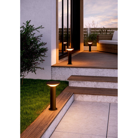Rigosa LED outdoor post light in black steel with a round white diffuser disc, shown lining steps and a garden path beside a wooden deck at dusk.