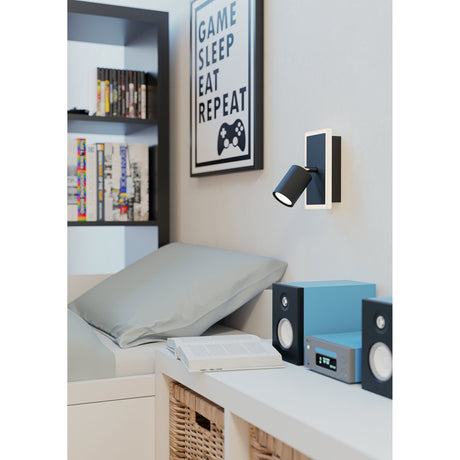Rimbocchi 1 LED wall spotlight in black steel with white-edged rectangular backplate and adjustable cylindrical head, mounted above a bedside desk in a bedroom setting.