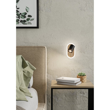 Rimbocchi LED wall spotlight in black and brown, oval wood-grain plate with white surround and adjustable black cylindrical head, mounted beside a bedside table in a bedroom setting.