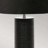 A close-up of the Ripple 1 Light Table Lamp in black, showcasing its cylindrical textured base with horizontal grooves and a large lampshade against a neutral backdrop. This contemporary lighting piece boasts a minimalist design.