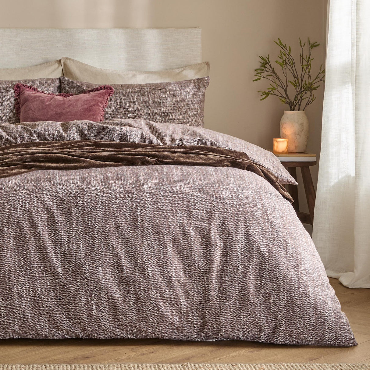 Harris Cotton Herringbone Duvet Cover Set in Chocolate on a bed with cream headboard, pink velvet cushions and a brown throw, styled in a bedroom with a bedside stool and vase.