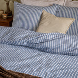 Holme Stripe Cotton Brushed Duvet Cover Set in denim blue and white, shown on a bed with matching striped pillowcases and a plain beige cushion, in a bedroom setting.
