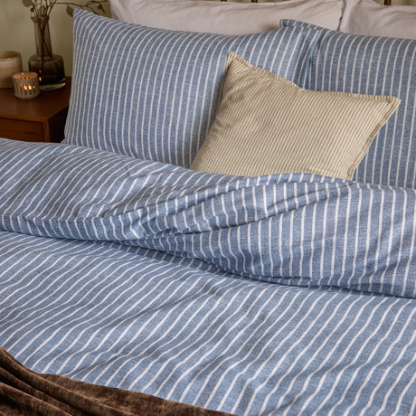 Holme Stripe Cotton Brushed Duvet Cover Set in denim blue and white, shown on a bed with matching striped pillowcases and a plain beige cushion, in a bedroom setting.