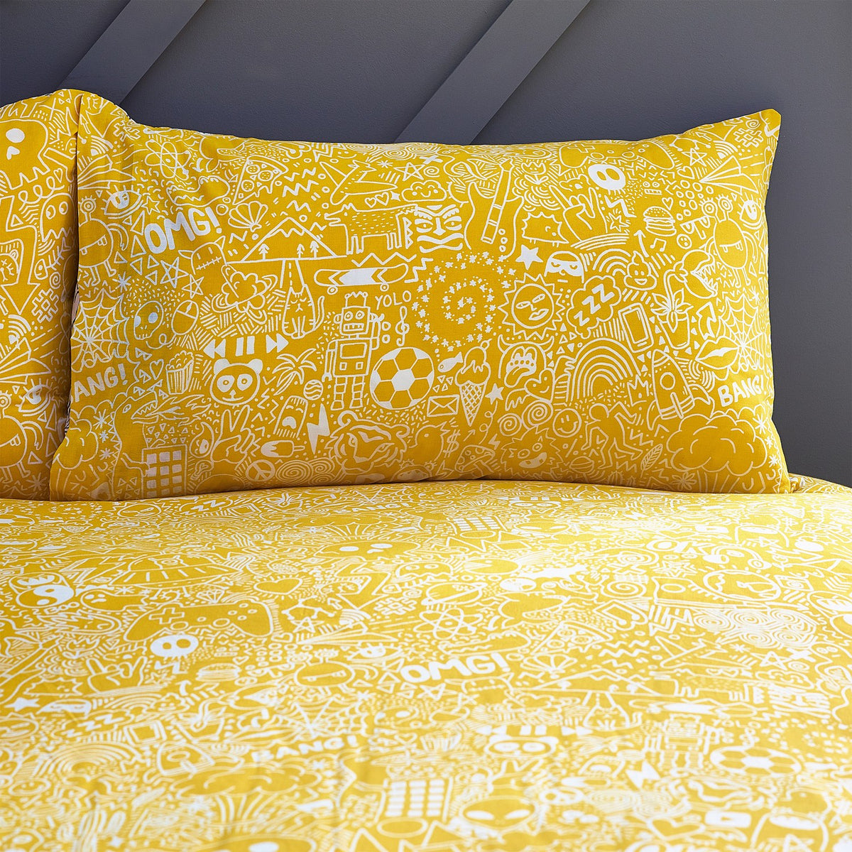 Doodles Polycotton Printed Duvet Cover Set on a bed, yellow reverse side shown with white doodle print of characters, symbols and speech-bubble words, matching pillowcase against a grey wall.