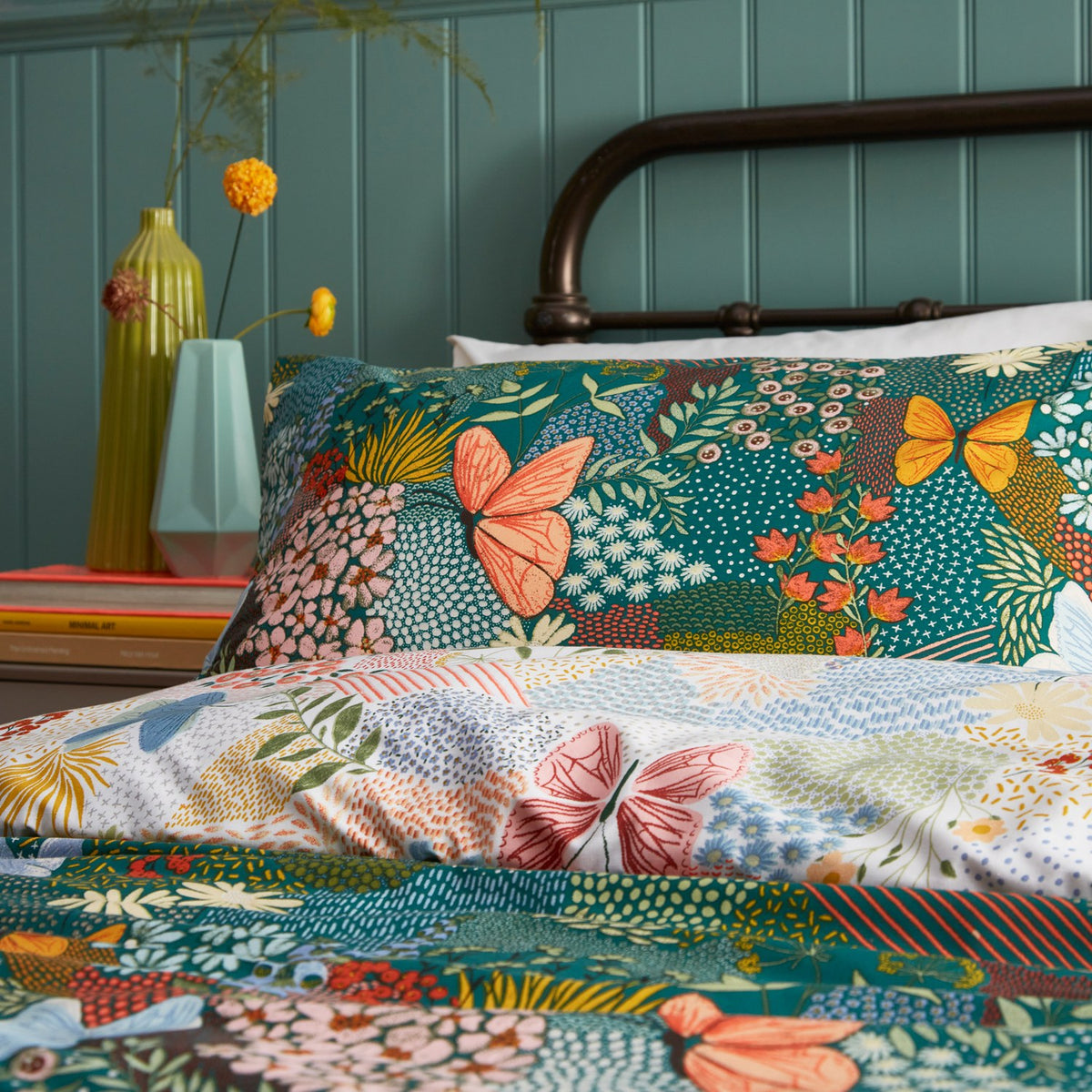 Forage Printed Floral Duvet Cover Set in teal, styled on a bed with a metal frame, showing coral, blue and amber butterflies among a patchwork floral print.