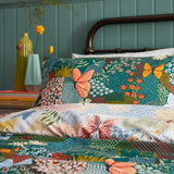 Forage Printed Floral Duvet Cover Set in teal, styled on a bed with a metal frame, showing coral, blue and amber butterflies among a patchwork floral print.