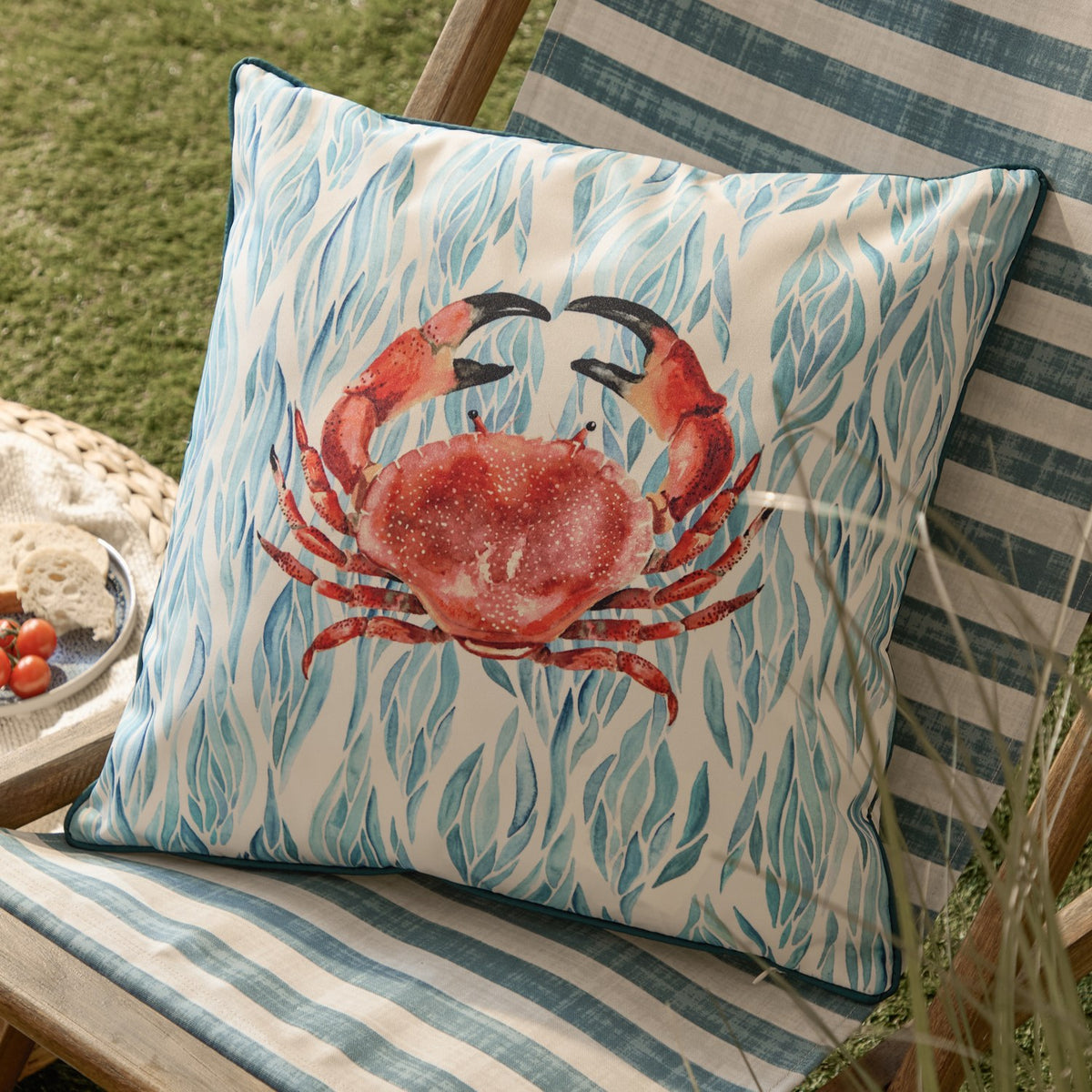 Abersoch Abstract polyester cushion in Marine, 45cm square with a coral-red crab design on a blue and teal seaweed print, shown on a striped garden chair.