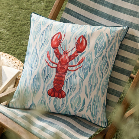 Abersoch Abstract Polyester Cushion in Marine, square with teal piped edge, showing a red lobster on a cream ground with blue-green leaf shapes, styled on a striped garden chair.