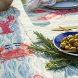 Abersoch Abstract Polyester Placemats in Marine, showing crab and lobster motifs on a blue and coral background, styled with rosemary sprigs and a blue bowl of olives.