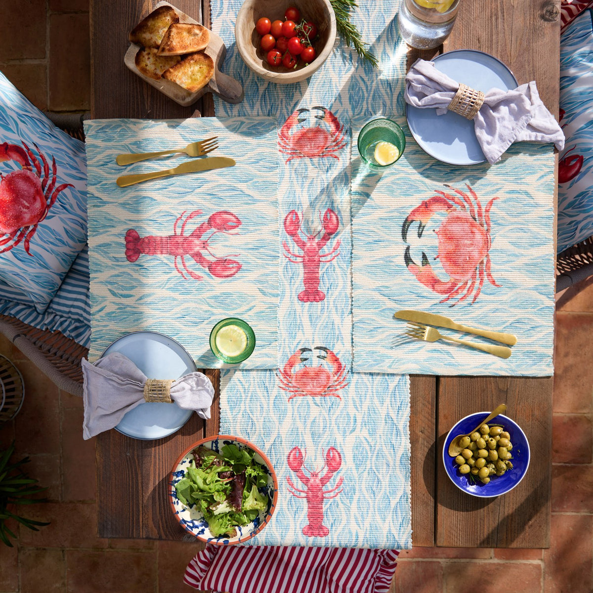 Abersoch Abstract Polyester Placemats in Marine, blue and coral fabric mats with crab and lobster prints, styled on an outdoor wooden table with plates, cutlery, and food.