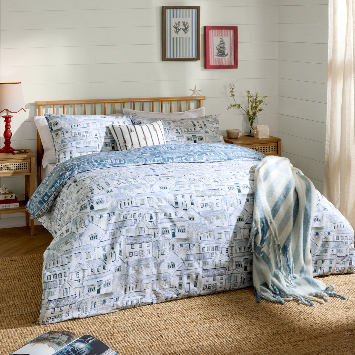 Bedroom featuring the Abersoch Town Cotton Reversible Duvet Cover Set in Sea Blue with hand-painted coastal print, matching pillowsham, and striped throw on a wooden bed frame.