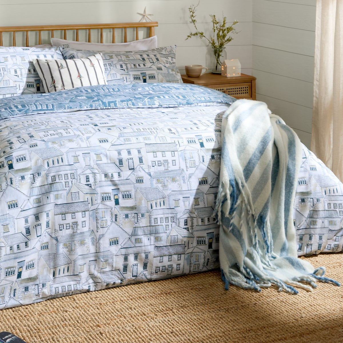 Abersoch Town Cotton Reversible Duvet Cover Set in Sea Blue on a bed, showing a hand-drawn coastal houses print in cream, blue and grey, with striped cushions and a blue striped throw.