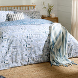 Abersoch Town Cotton Reversible Duvet Cover Set in Sea Blue on a bed, showing a hand-drawn coastal houses print in cream, blue and grey, with striped cushions and a blue striped throw.