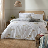 Abersoch Shore cotton stripe duvet set in Sky Blue on a bed with cane headboard, showing sailboat print and reverse stripe sides, styled with striped cushions in a neutral bedroom.