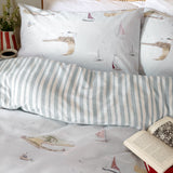 Abersoch Shore cotton duvet cover set in sky blue, showing sailboat and coastal print pillowcases alongside the reverse sky blue and white stripe duvet, styled on a bed with a book.