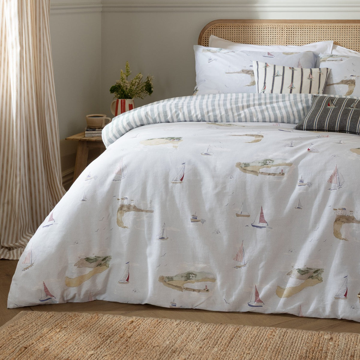 Abersoch Shore Cotton Stripe Duvet Cover Set in Sky Blue, dressed on a bed with sailboat and coastal island print, striped reverse, and matching cushions in a bedroom setting.