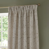 Acanthus Jacquard Linen pencil pleat curtains in beige with a woven ivory-cream leaf pattern, shown pleated on a brass pole beside a window.