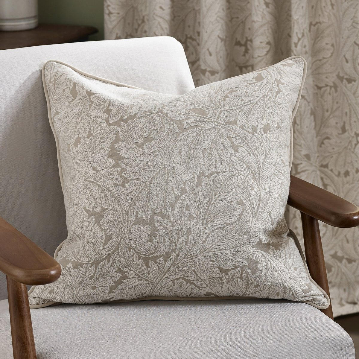 Acanthus Jacquard Linen Cushion in cream, a square cushion with woven acanthus leaf pattern and piped edge, shown on a light upholstered armchair.