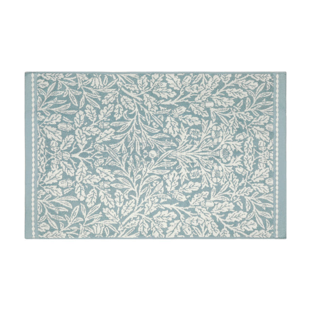 Rectangular cotton bath mat in mineral blue with an all-over off-white Acorn floral and foliage pattern, framed by slim beaded side borders, on a plain white background.