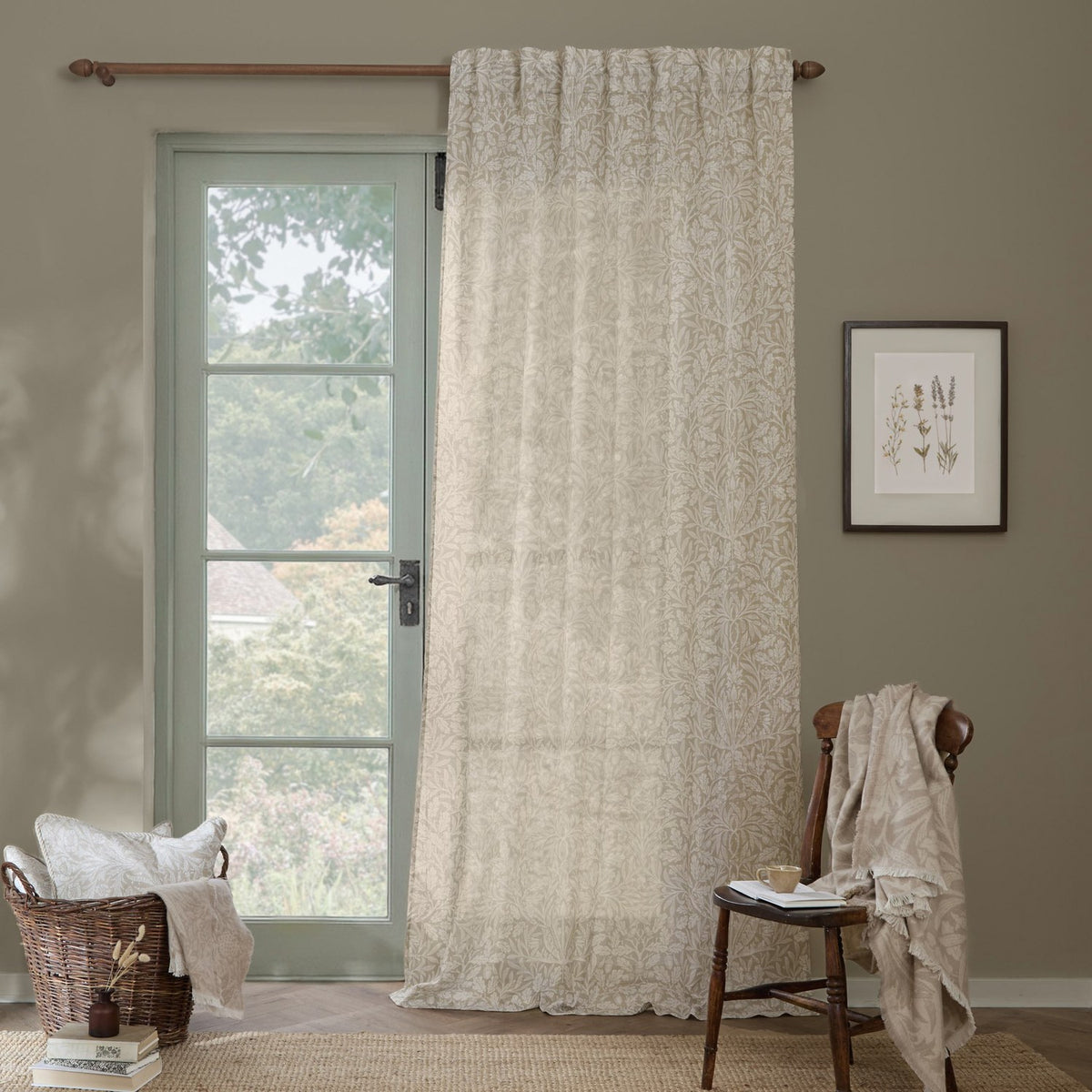 Acorn Damask Linen Curtain Panel in natural cream, hung from a wooden pole beside a glazed door in a living room with a chair, basket, and framed botanical print.