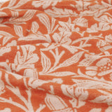 Close-up of the Acorn Tonal Jacquard Cotton Blend Throw in brick orange, showing the woven cream botanical pattern of leaves and acorns across softly draped fabric folds.