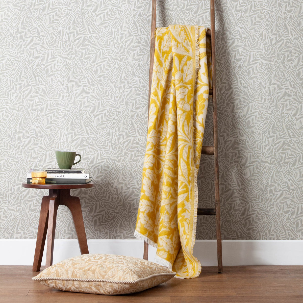 Acorn Tonal Jacquard Cotton Blend Throw in gold and cream draped over a wooden ladder, with a matching cushion, side table and patterned wallpaper in the room setting.