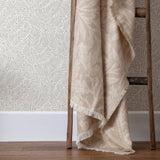 Acorn Tonal Jacquard Linen Throw in beige draped over a wooden ladder, showing fringed edges and a woven leaf pattern against botanical-print wallpaper.