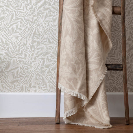 Acorn Tonal Jacquard Linen Throw in beige draped over a wooden ladder, showing fringed edges and a woven leaf pattern against botanical-print wallpaper.