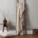 Acorn Tonal Jacquard Linen Throw in beige draped over a wooden ladder, showing its fringed edge and leafy woven pattern in a living room setting.